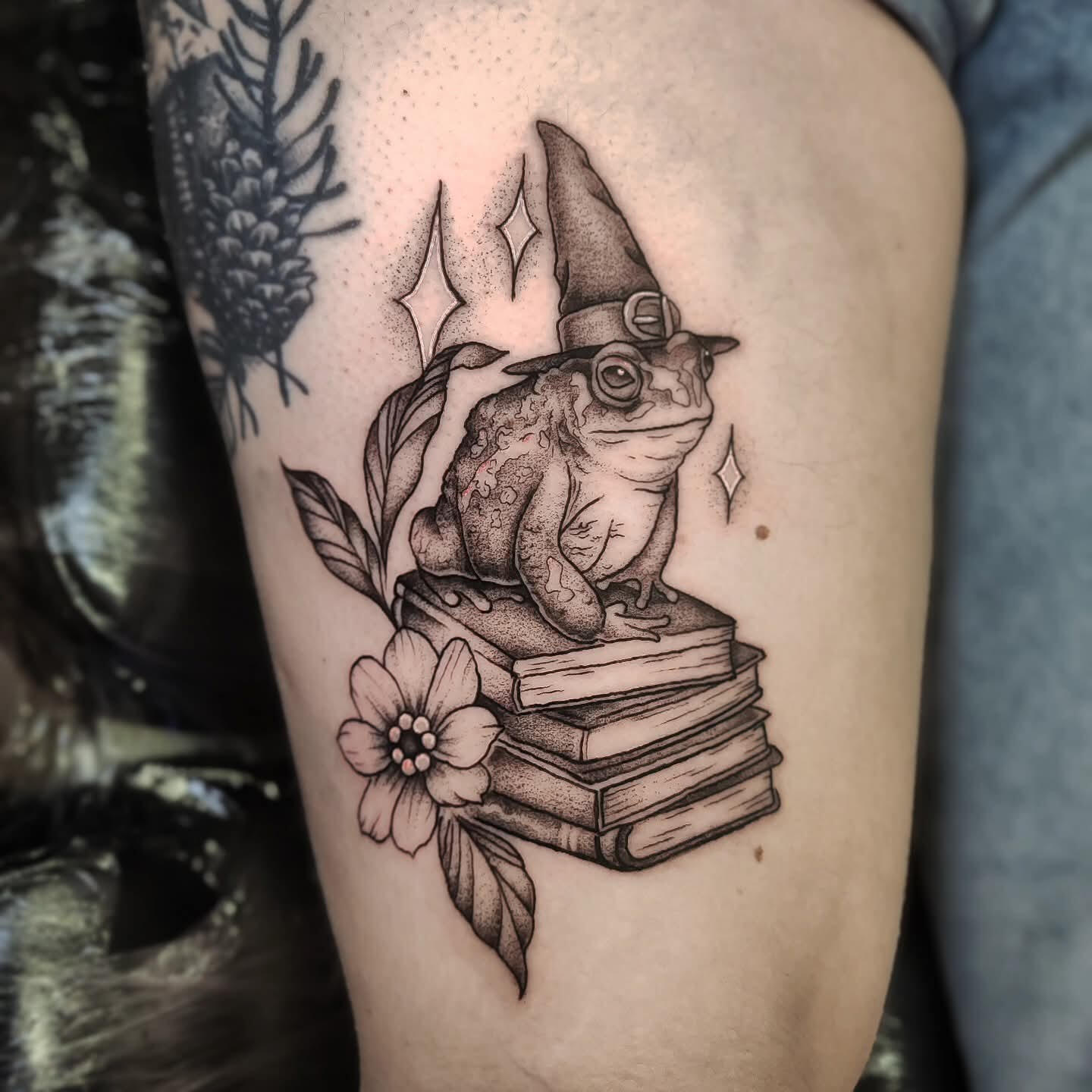Blackwork tattoo of a toad wearing witch hat sitting on a pile of old books